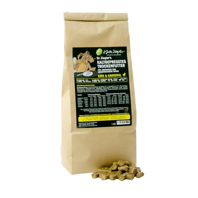 Braune Papiertüte mit schwarzem Etikett, beschriftet mit 'Dr. Ziegler's KALTGEPRÄSSTES TROCKENFUTTER', enthält Trockenfutter für Hunde mit Enten- und Kartoffelanteil. Vor der Tüte liegen einige Futterpellets.