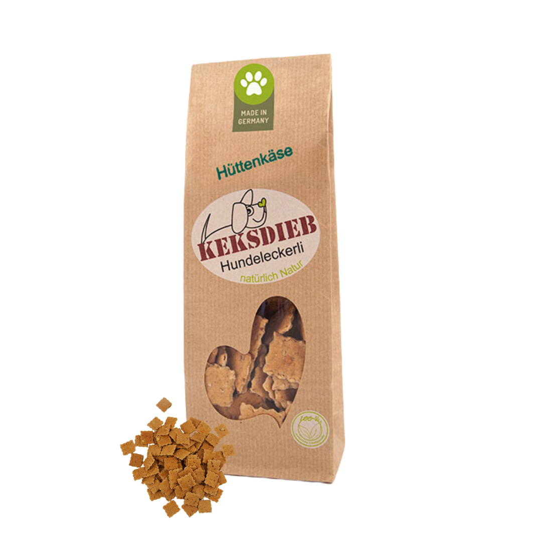 Papiertüte mit der Aufschrift 'Keksdieb Hundeleckerli Hüttenkäse', gefüllt mit quadratförmigen Hundekeksen, die vor der Tüte liegen.