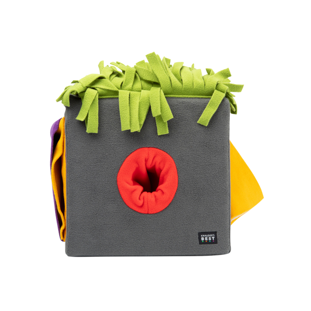 Graue Futterbox mit bunten, fransigen Stoffstreifen in Grün, Orange und Gelb. Die Box hat eine runde Öffnung in der Mitte.