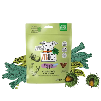 Grüne Verpackung mit der Aufschrift 'VEGDOG Veggies Snack', Bild eines weißen Hundes mit Karotte im Maul, umgeben von Illustrationen von Kastanien und grünen Blättern.