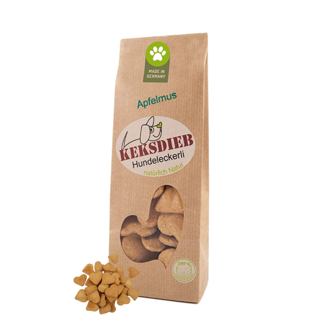 Papiertüte mit der Aufschrift 'Keksdieb Hundeleckerli Apfelmus', gefüllt mit herzförmigen Hundekeksen, die vor der Tüte liegen.