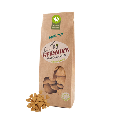 Papiertüte mit der Aufschrift 'Keksdieb Hundeleckerli Apfelmus', gefüllt mit herzförmigen Hundekeksen, die vor der Tüte liegen.
