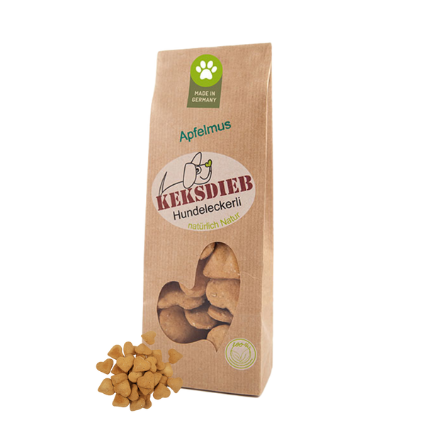 Papiertüte mit der Aufschrift 'Keksdieb Hundeleckerli Apfelmus', gefüllt mit herzförmigen Hundekeksen, die vor der Tüte liegen.