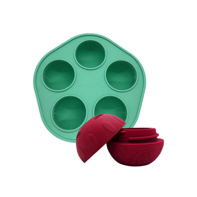 Grüne Silikonform mit fünf runden Vertiefungen und zwei rote, halbkugelförmige Hundespielzeuge mit Muster und Schraubverschluss.